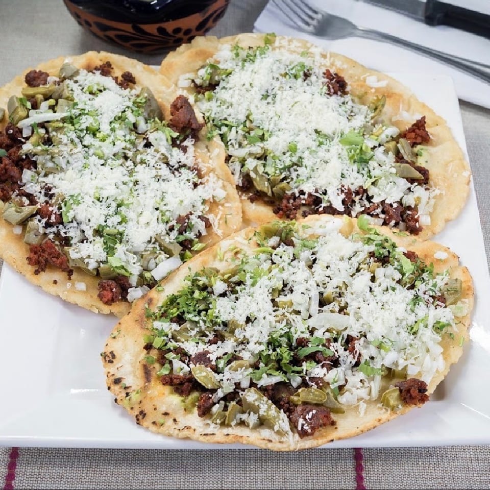Three Mexican sopes with chorizo, nopales, onions, cilantro, and crumbled cheese on a white plate.