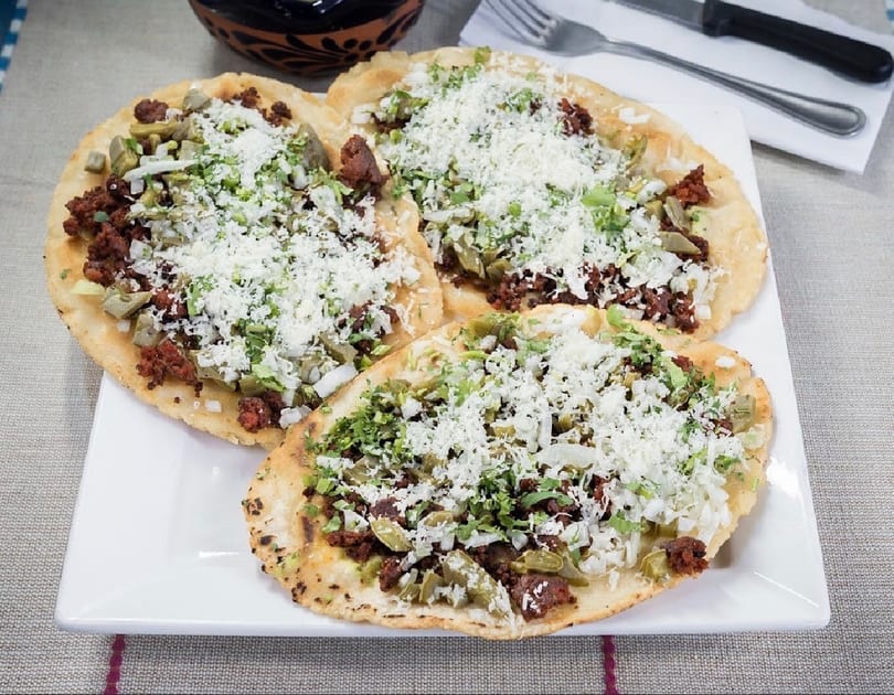 Three Mexican sopes with chorizo, nopales, onions, cilantro, and crumbled cheese on a white plate.