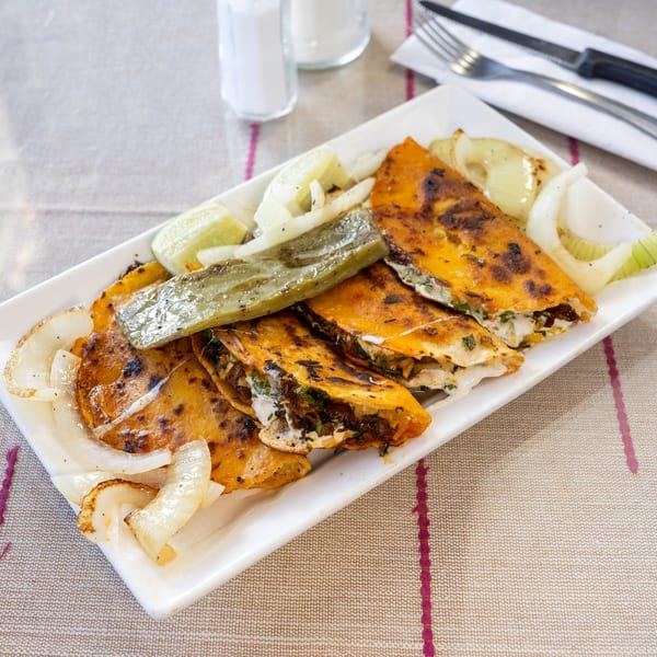 A white rectangular plate filled with golden-brown quesadillas, grilled onions, lime wedges, and a green chili.