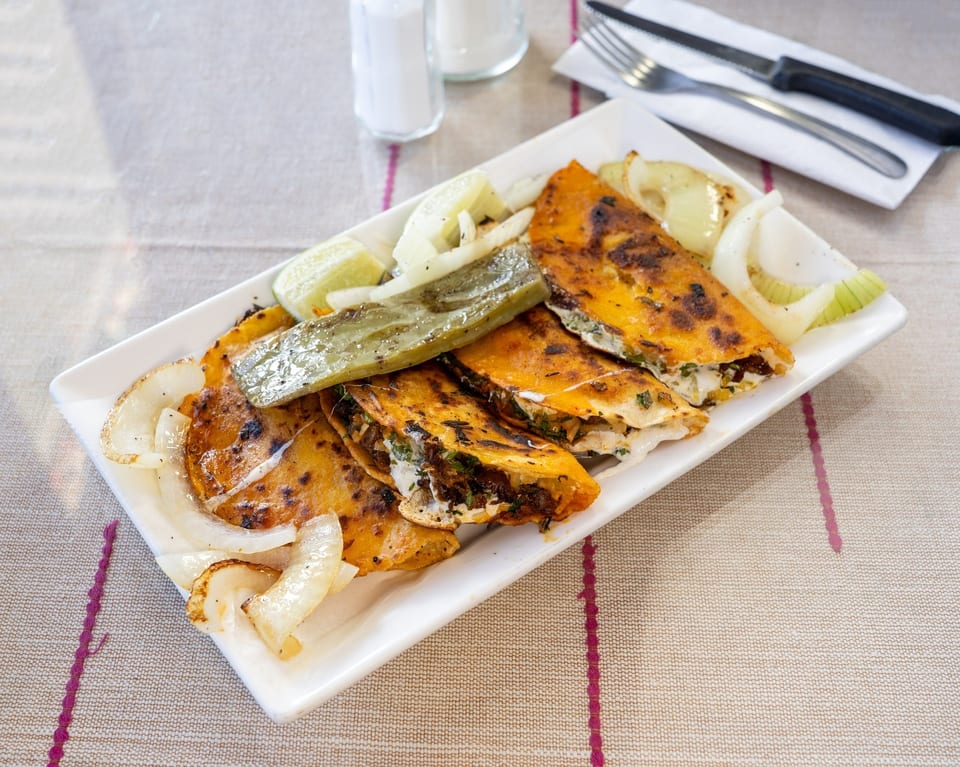 A white rectangular plate filled with golden-brown quesadillas, grilled onions, lime wedges, and a green chili.