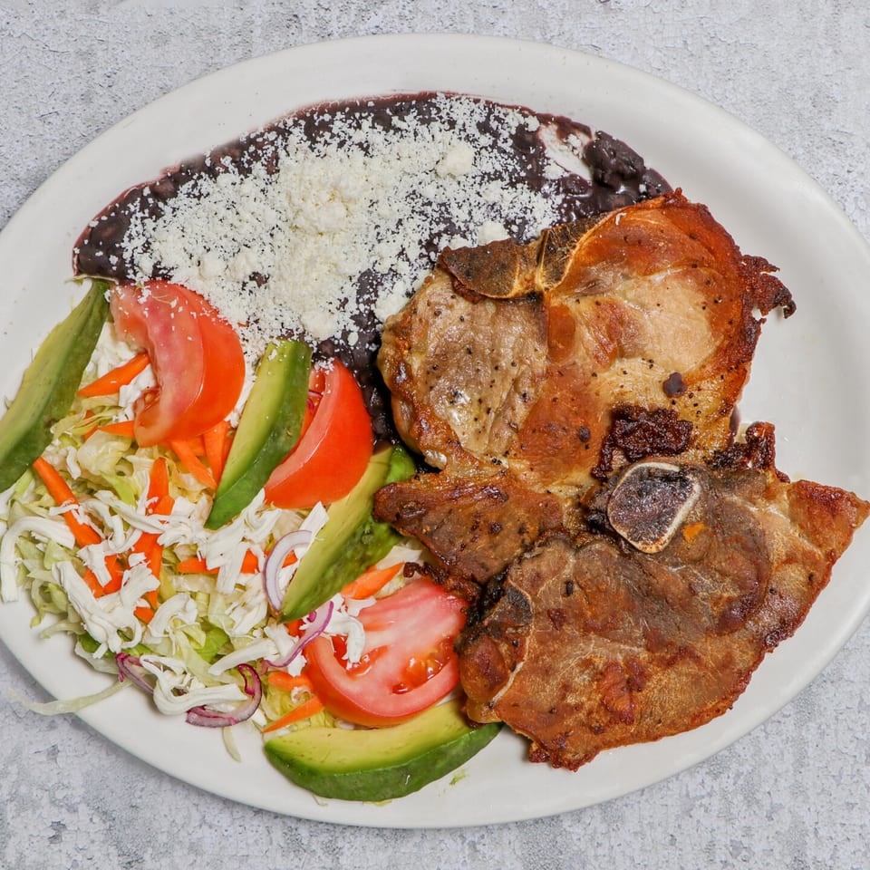 Two grilled pork chops with black beans, shredded cheese, and a side salad with avocado and tomato.