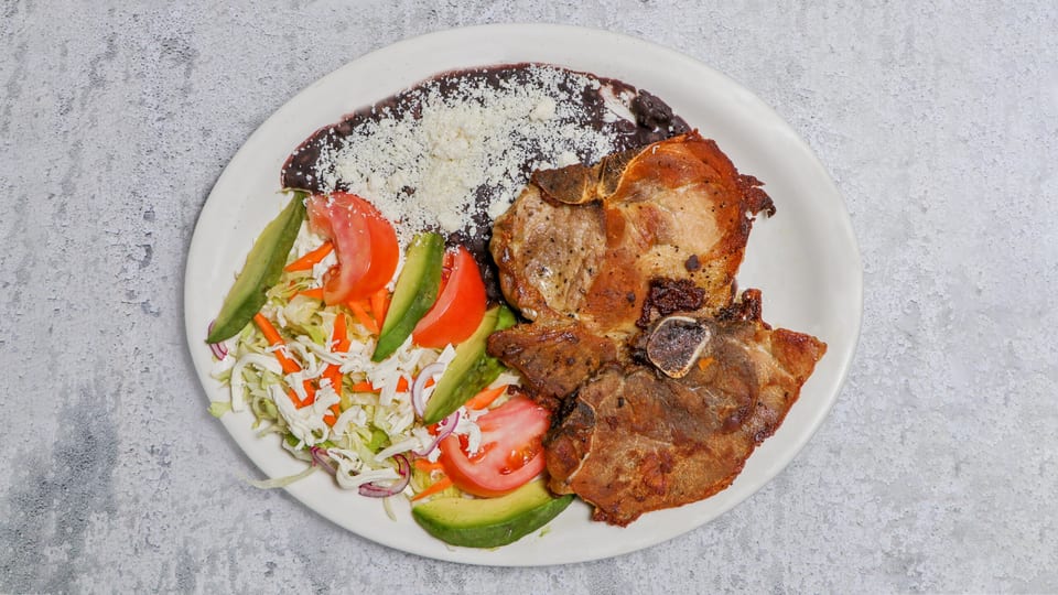 Two grilled pork chops with black beans, shredded cheese, and a side salad with avocado and tomato.