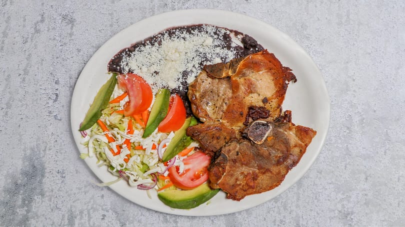 Two grilled pork chops with black beans, shredded cheese, and a fresh salad with avocado and tomato.