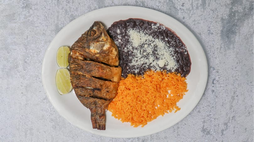 Fried fish with lime wedges, black beans topped with cheese, and orange rice on a white plate.