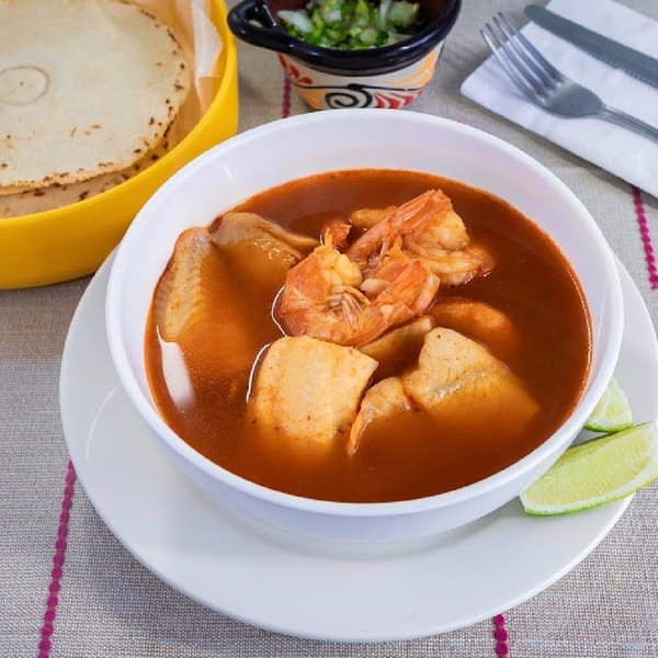 Seafood soup with shrimp, fish, and lime wedges, served with tortillas and chopped onions.