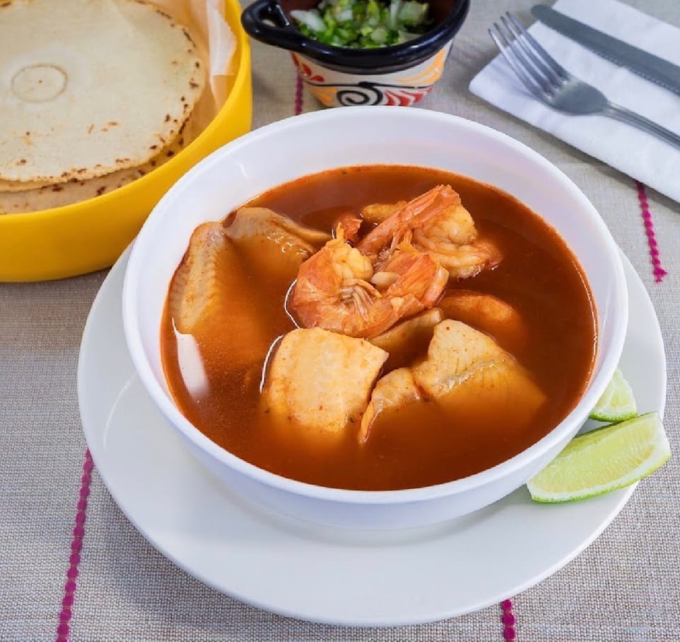 Seafood soup with shrimp, fish, and lime wedges, served with tortillas and chopped onions.